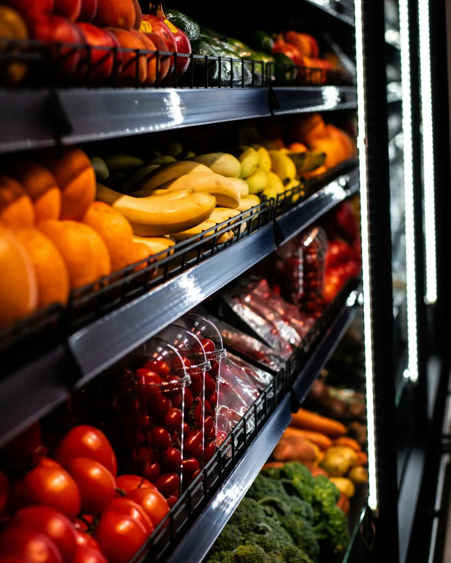 Tiered supermarket shelves of fruit and vegetables receding into depth.
